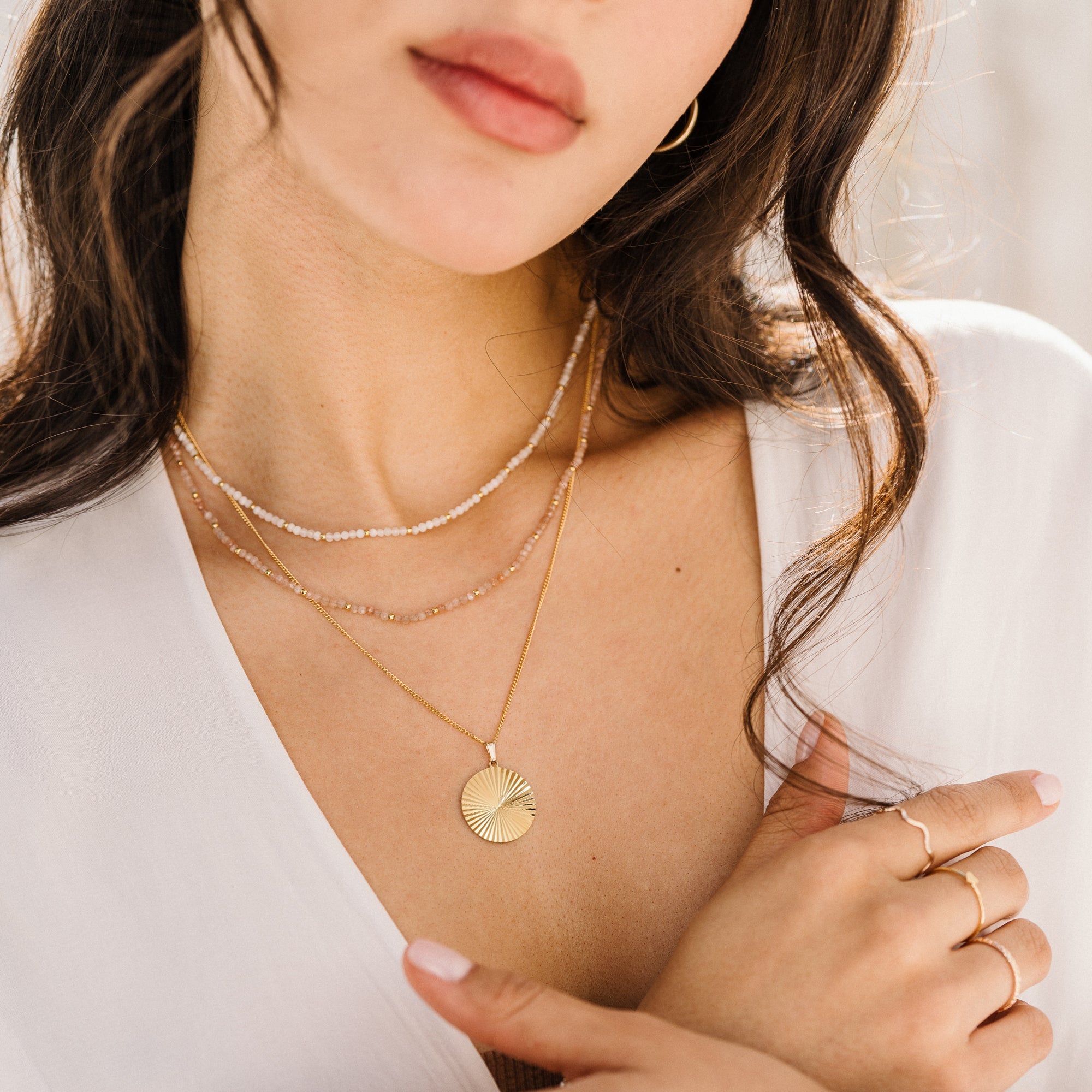 Close-up of a woman’s chest wearing the Sunburst Pendant Necklace and two other necklaces. She also has her hands closed and is wearing three rings.