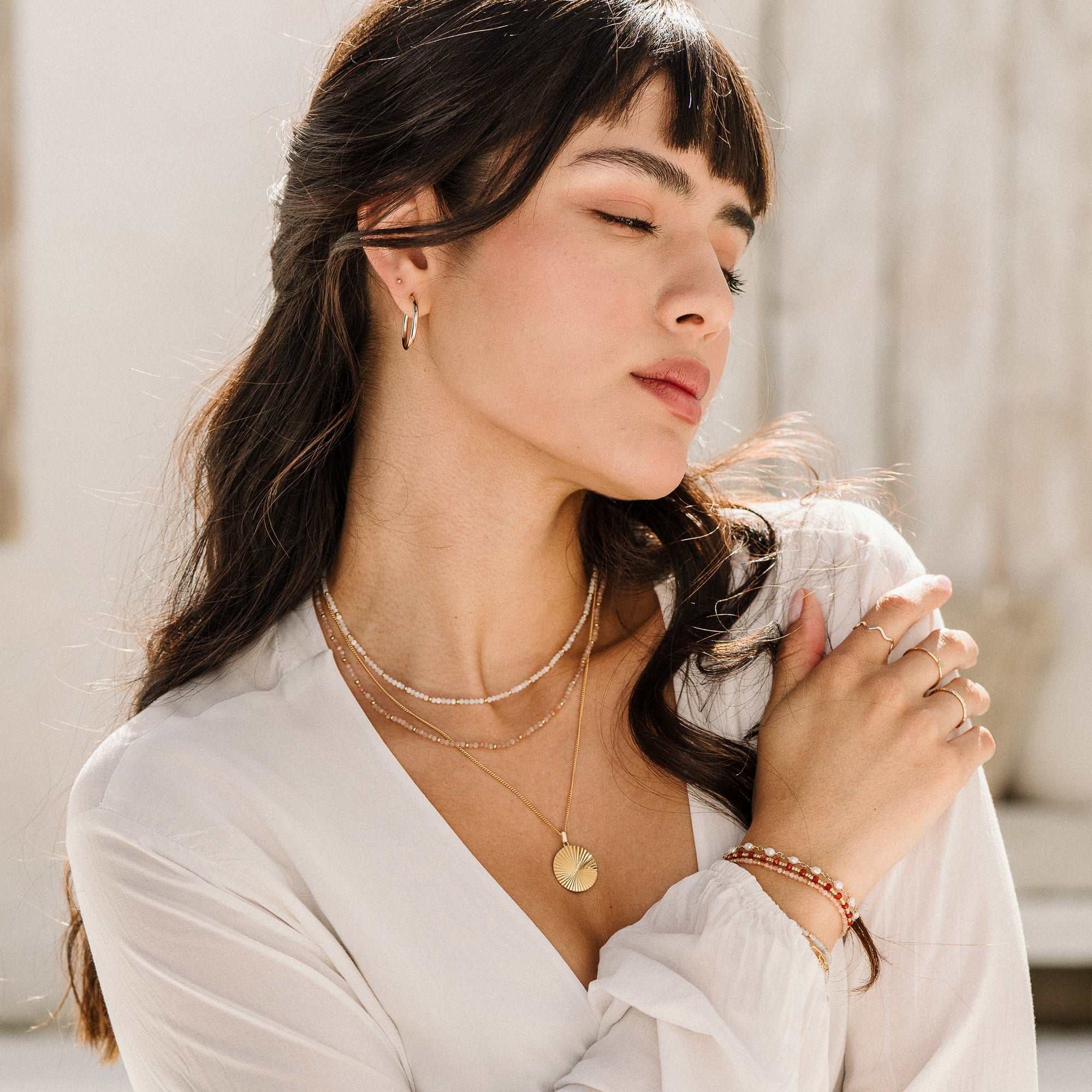 Woman wearing three necklaces, including the Sunburst Pendant Necklace, styled with a beige cover and a brown top. She is wearing bracelets, rings, and hoop earrings. 