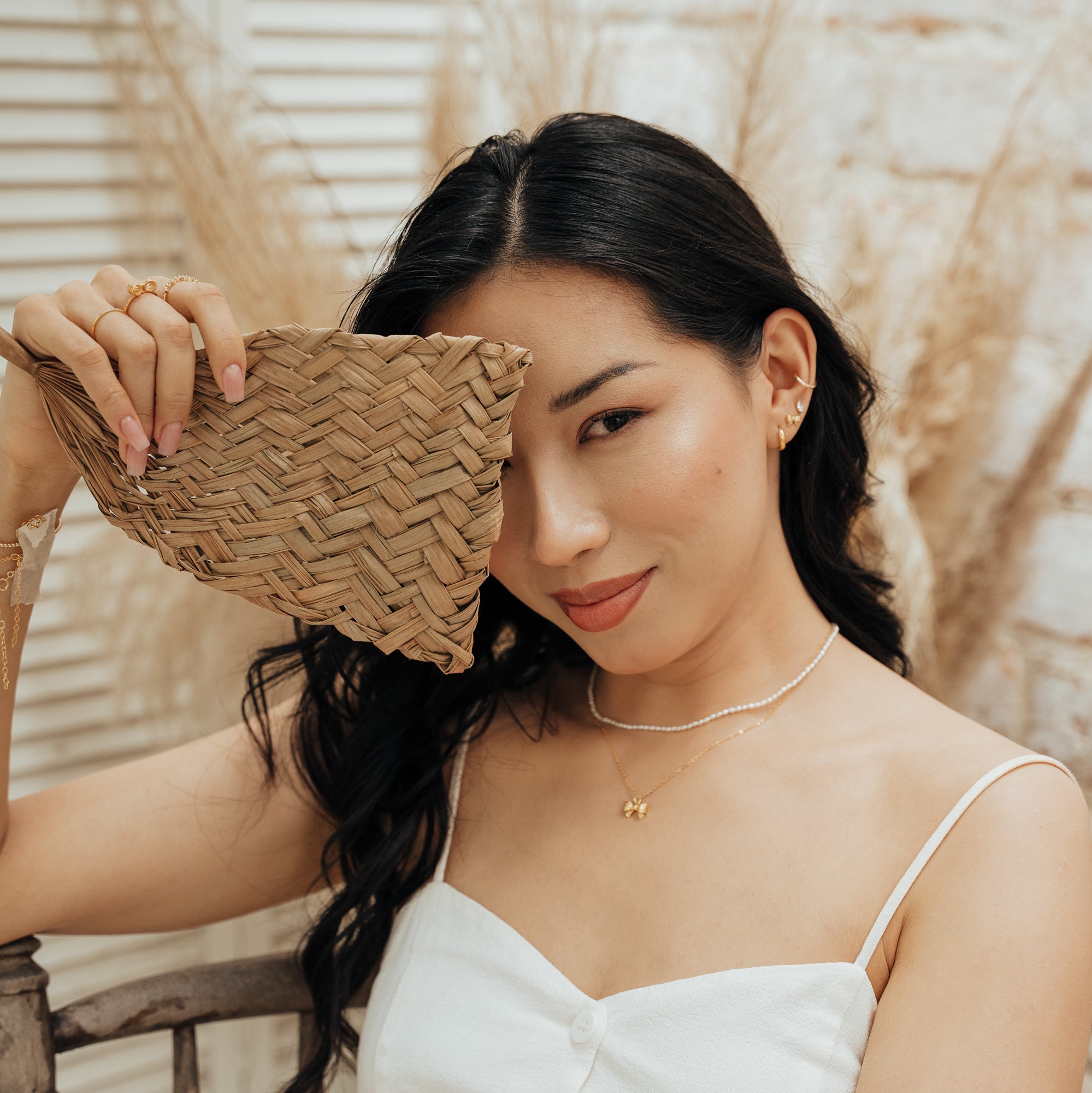 Woman covering one eye with a raffia fan. She is wearing the Dainty Pearl Strand Necklace and the Ribbon Bow Necklace around her neck. On her ears, she has a set of earrings and an ear cuff. On her right hand, she is wearing the Ribbon Bow Ring on her middle finger, the Alternating Diamond Ring on her index finger, and the Simple Stacking Ring on her ring finger.