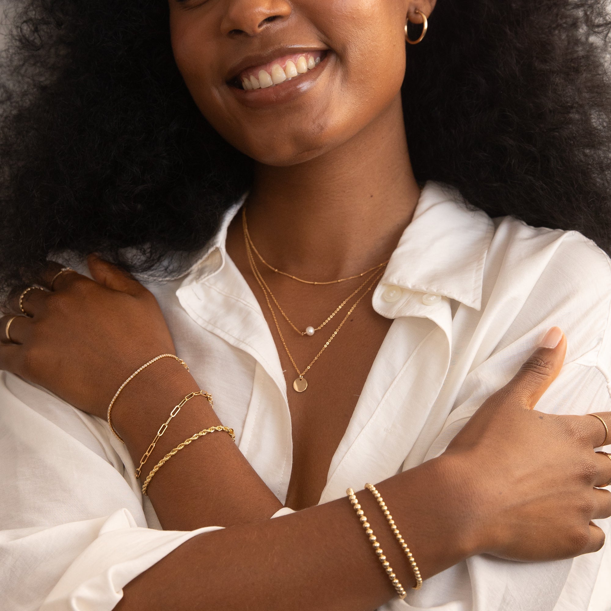 Woman wearing the Satellite & Coin Necklace Set and Freshwater Pearl Necklace, along with gold bracelets: Dainty CZ Tennis Bracelet, and Chunky Rope Chain Bracelet on the left wrist, and a Stretch Bead Bracelets on the right. She is dressed in a white button-up shirt, smiling with arms crossed to showcase the jewelry. Small Large Freshwater Pearl Necklace by Simple & Dainty Jewelry