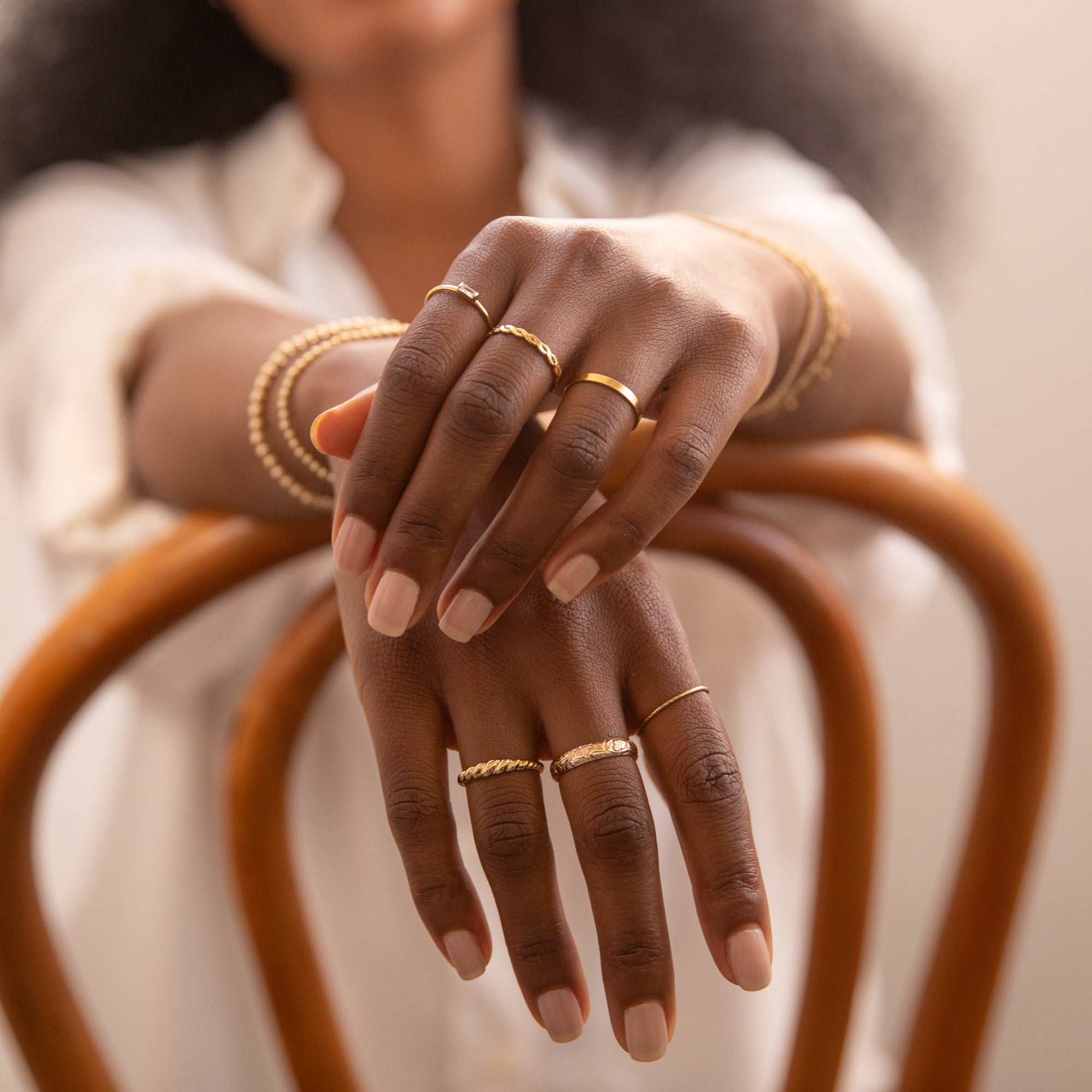 Close-up of hands resting on a wooden chair, wearing six gold rings—Dainty Baguette Ring, Twist Ring, Flat Band Ring, Croissant Ring, Thick Flower Band Ring, and Sparkle Stacking Ring—along with gold bracelets.