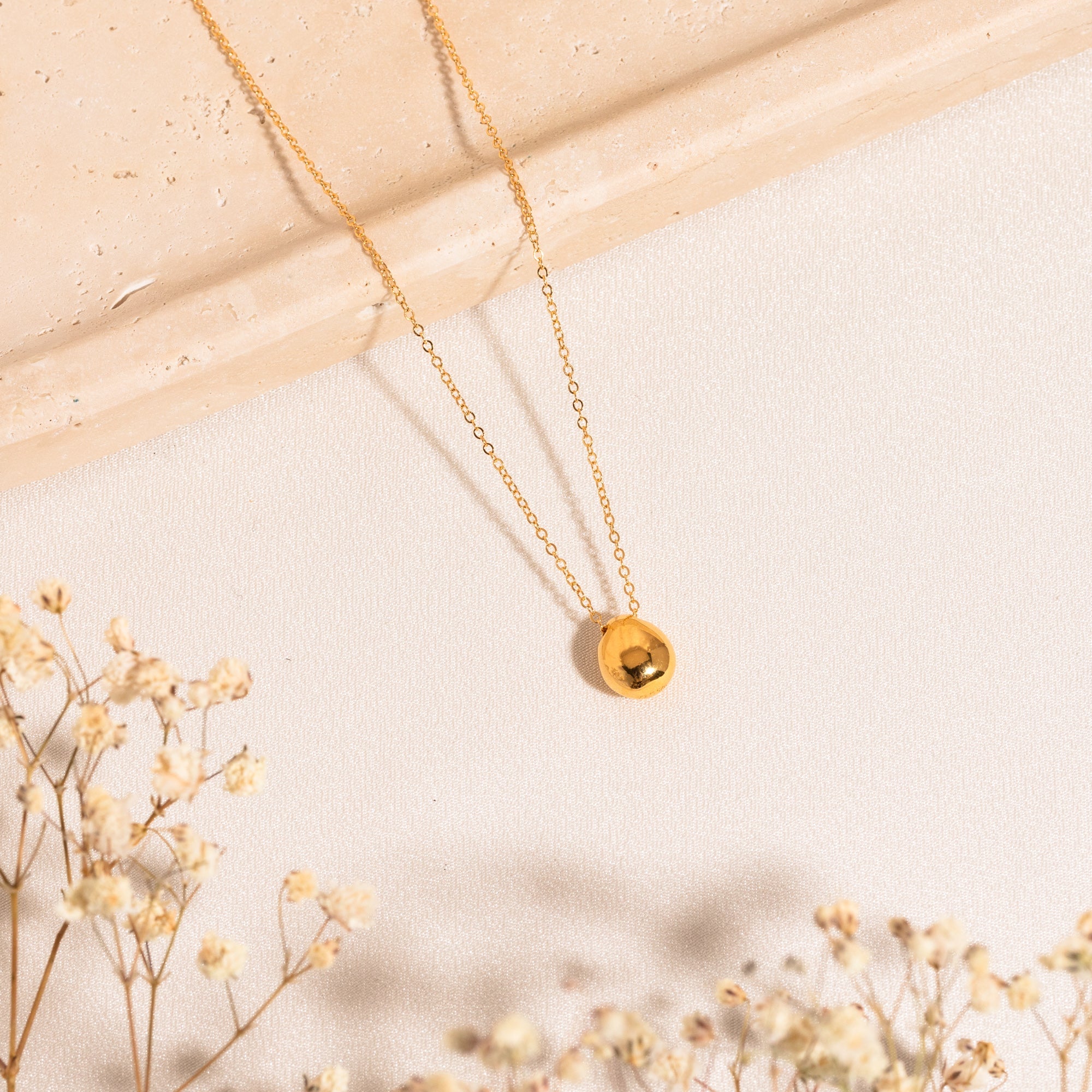 Pebble necklace lying on a white surface with sprigs of baby's breath; part of the chain rests on a marble surface.