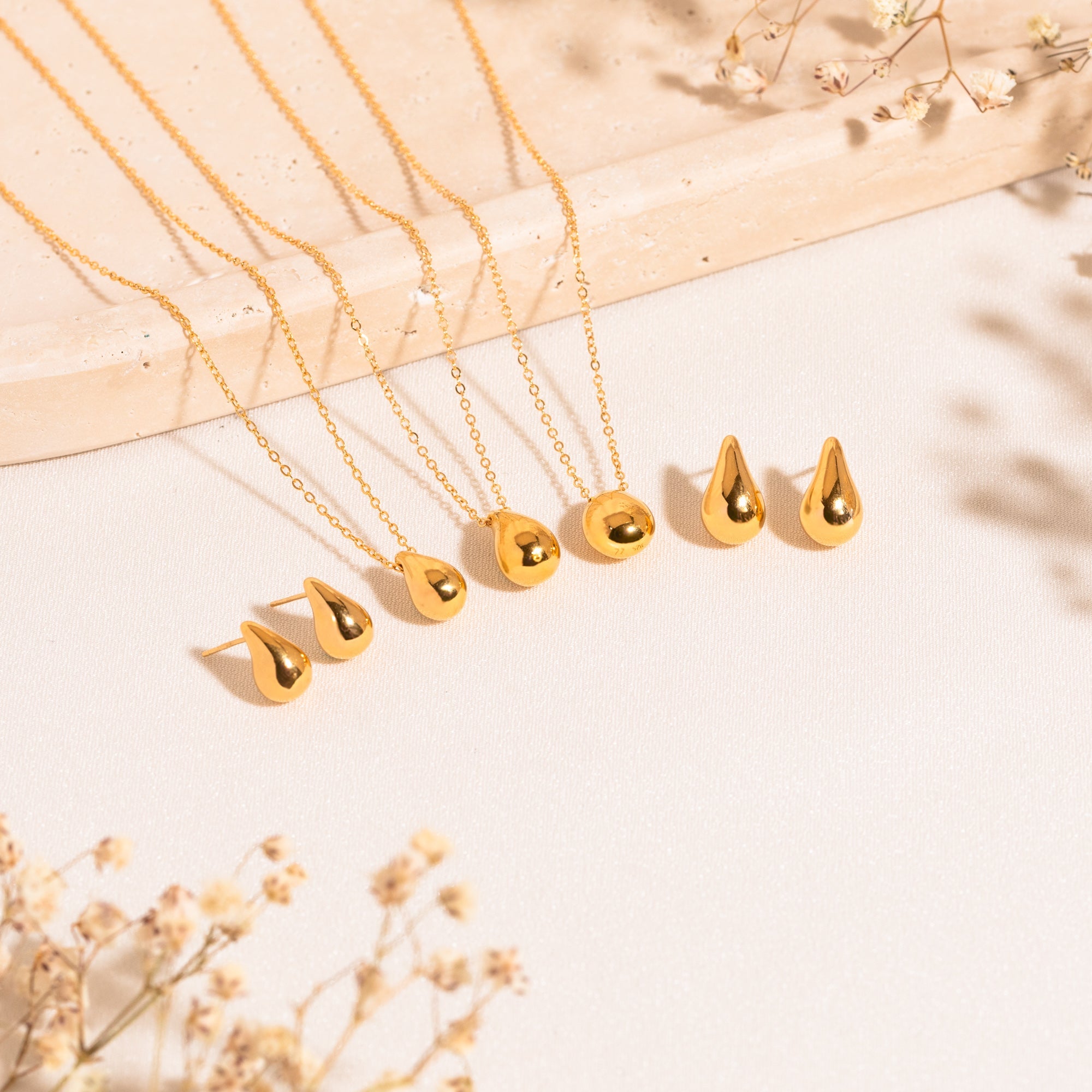 Different jewelry pieces lying on a white surface. From left to right: dainty teardrop earrings, dainty teardrop necklace, teardrop necklace, pebble necklace, and teardrop earrings. Baby's breath flowers are visible in the bottom-left corner of the image.