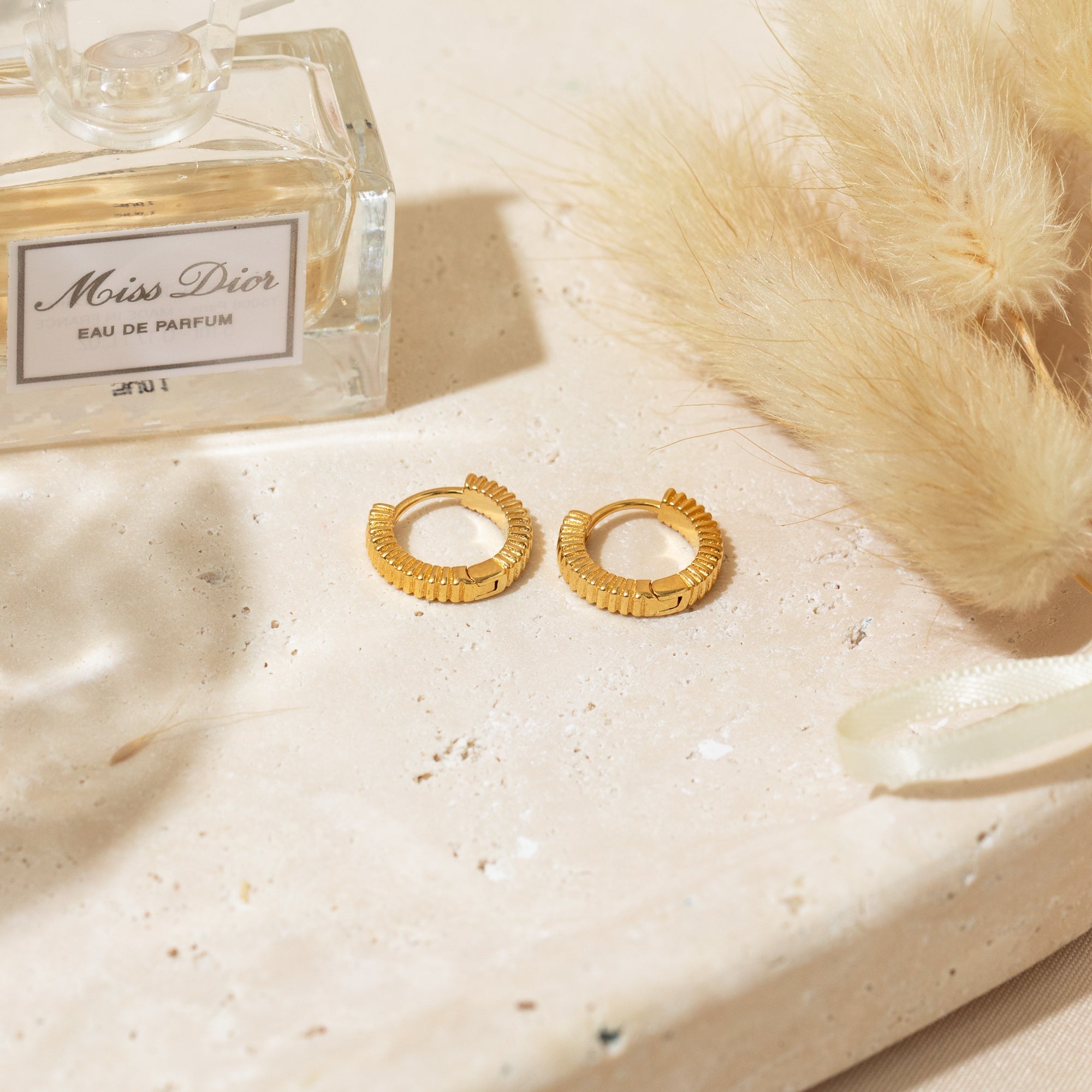 The Ribbed Huggie Hoop Earrings placed in a stone tray beside a perfume bottle and decorative feathers.