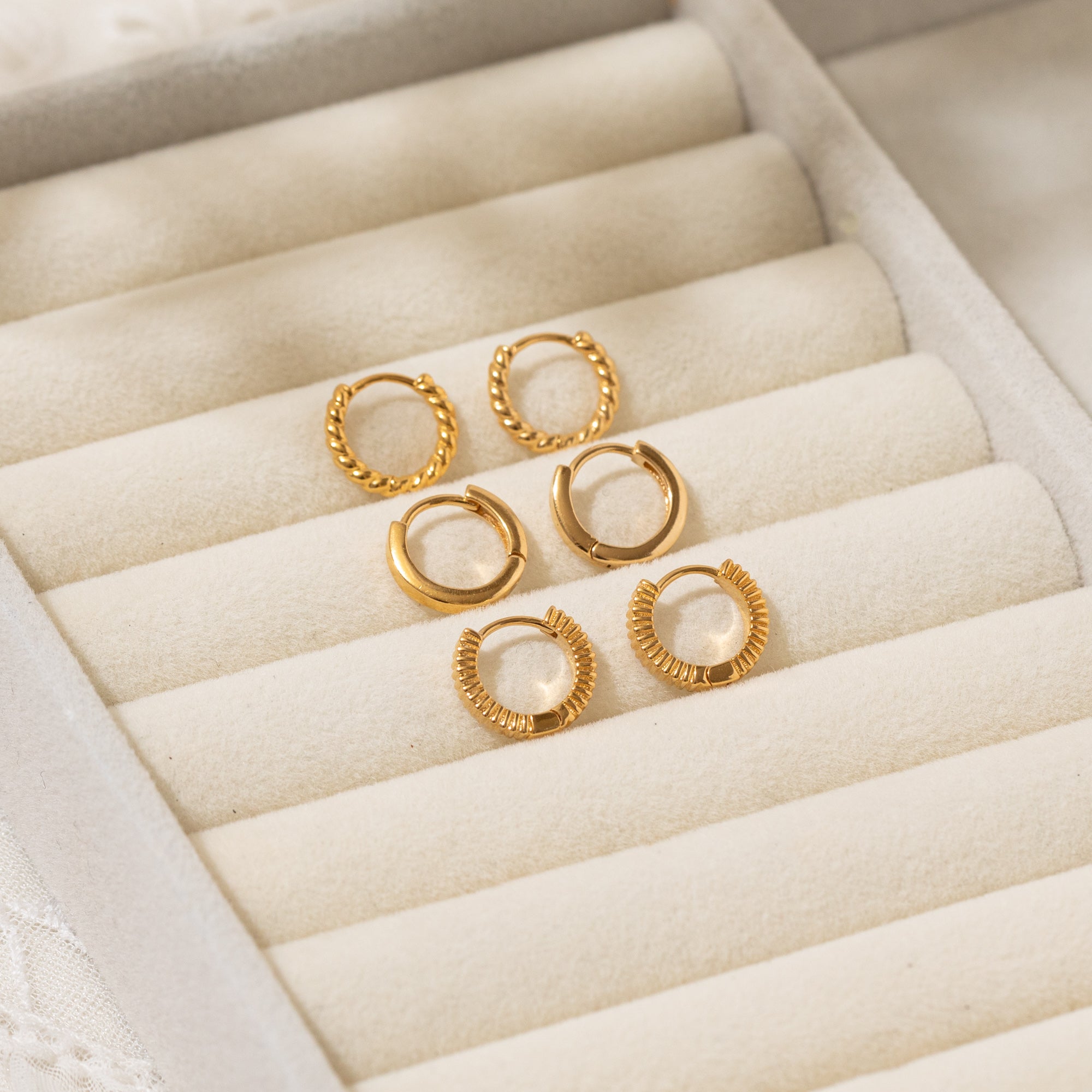 The Rope Huggie Hoop Earrings, Modern Huggie Hoop Earrings, and Ribbed Huggie Hoop Earrings placed in a beige jewelry box.