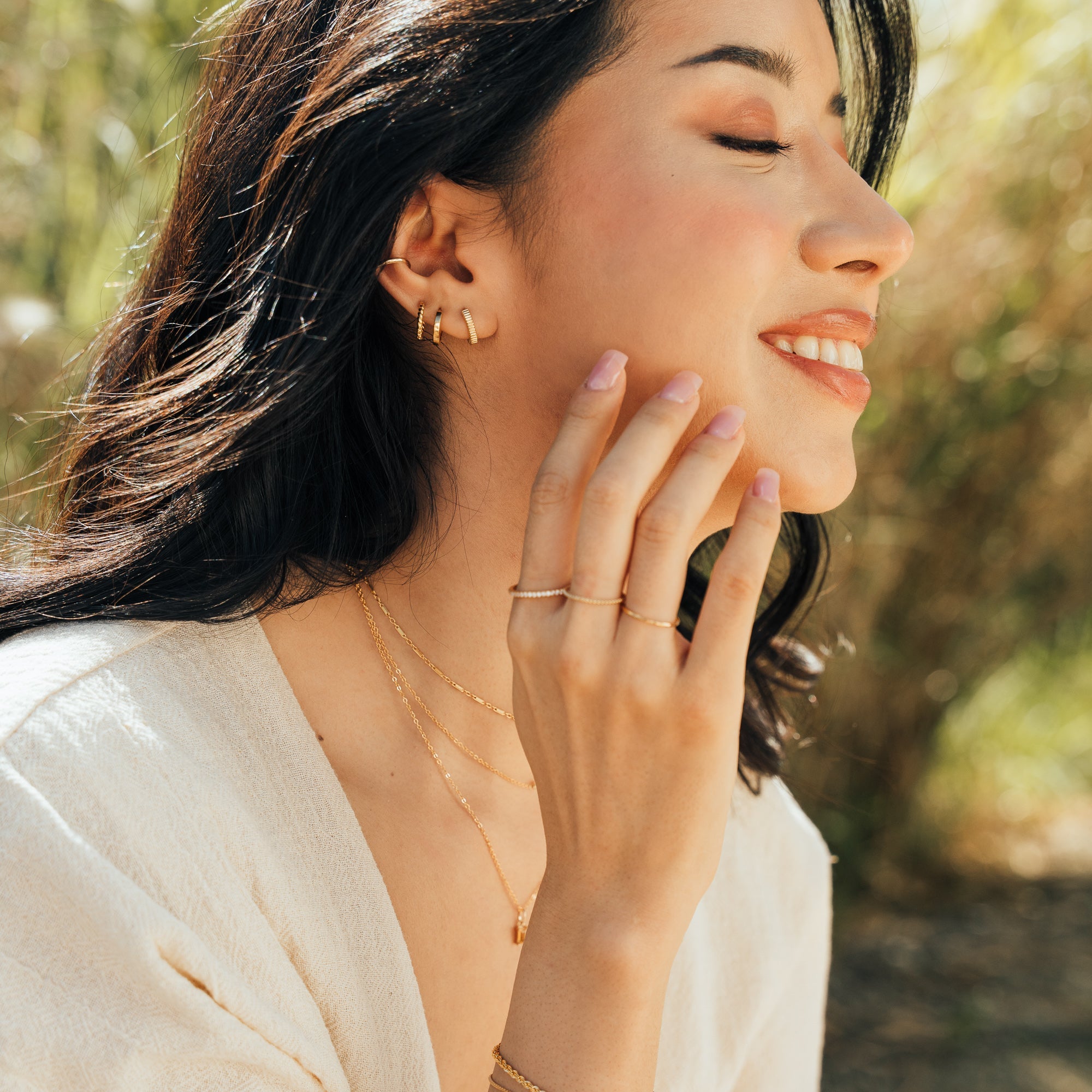 Woman wearing the Modern Huggie Hoop Earrings, Ribbed Huggie Hoop Earrings, Rope Huggie Hoop Earrings, and a Dainty Ear Cuff. She is touching her chin and wearing rings on three fingers and layered necklaces.